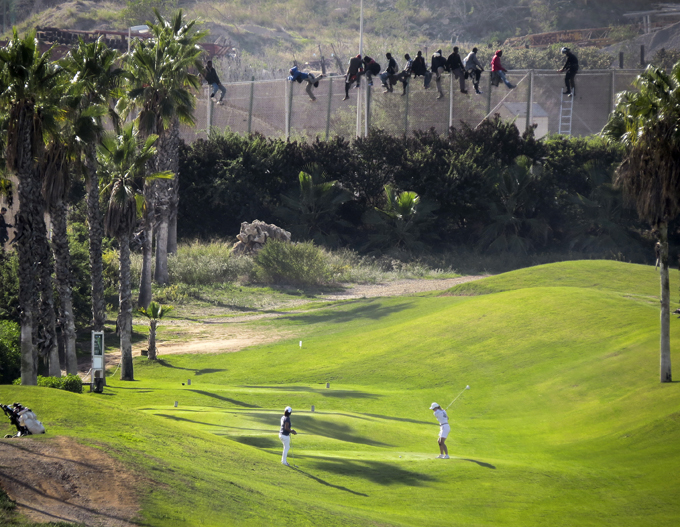 A golfer hits a tee shot as African migrants sit atop a border fence during an attempt to cross into Spanish territories between Morocco and Spain's north African enclave of Melilla, October 22, 2014. Photo by José Palazon/REUTERS.