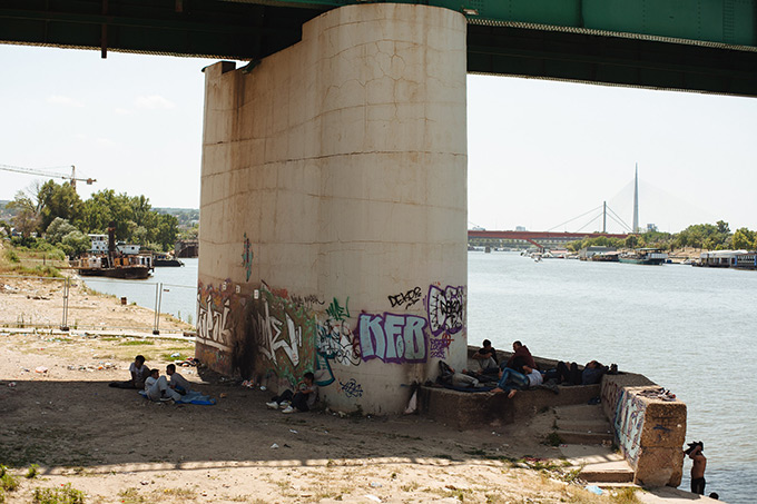 A group of migrants, under the bridge in Belgrade's Savamala district. The bridge provides shelter from the rain and shade from the strong midsummer sun. The area has since been closed off as a construction site, part of the often disputed "Belgrade Waterfront" project. July 2015. Author Luka Knežević Strika (Belgrade Raw).