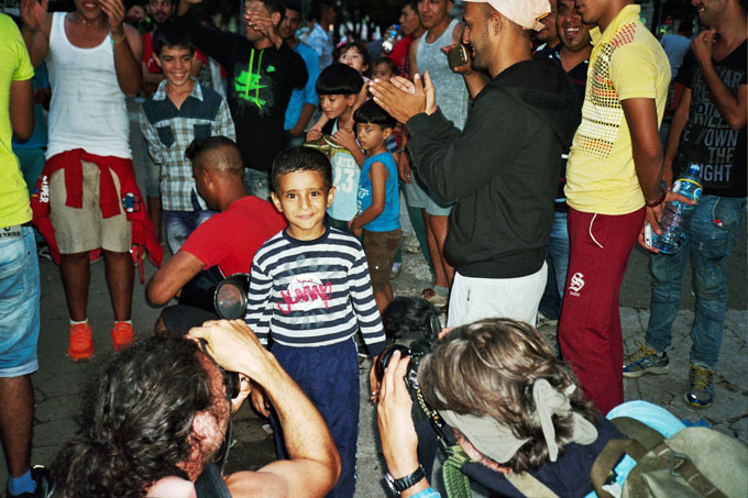 A migrant boy around six years old being photographed by photojournalists during the solidarity action, when the group No Borders makes tea for the refugees in the park. These actions happen every Saturday afternoon, and often they turn the park to a small party with traditional Syrian music and dancing.